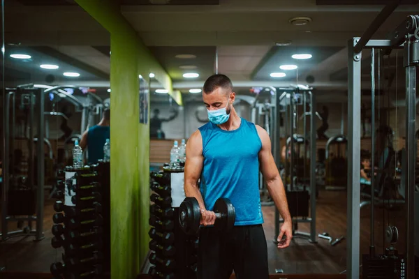Yüzünde maske olan beyaz bir sporcu spor salonunda ağırlık kaldırıyor ve egzersiz yapıyor. COVID 19 koronavirüs koruması