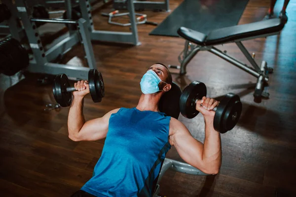 Yüzünde maske olan beyaz bir sporcu spor salonunda ağırlık kaldırıyor ve egzersiz yapıyor. COVID 19 koronavirüs koruması