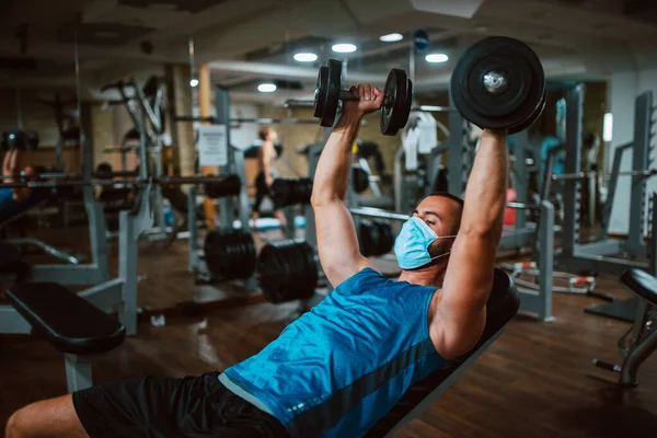 Yüzünde maske olan beyaz bir sporcu spor salonunda ağırlık kaldırıyor ve egzersiz yapıyor. COVID 19 koronavirüs koruması
