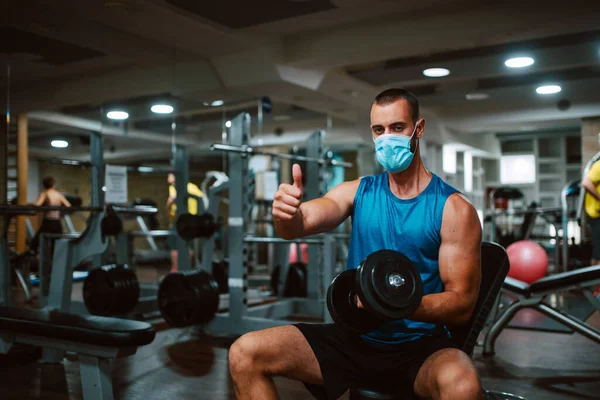 Yüzünde maske olan, elinde bir ağırlıkla genç kaslı beyaz bir sporcu spor salonunda baş parmağını kaldırıyor. COVID 'e karşı koruma - 19 koronavirüs