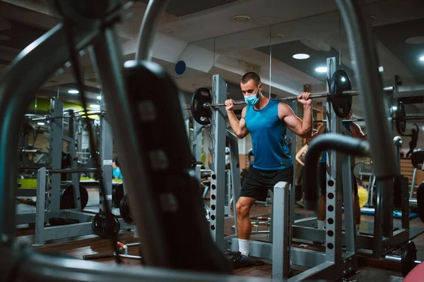 Yüzünde maske olan beyaz bir sporcu spor salonunda ağırlık kaldırıyor ve egzersiz yapıyor. COVID 19 koronavirüs koruması