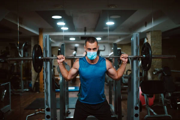 Yüzünde maske olan beyaz bir sporcu spor salonunda ağırlık kaldırıyor ve egzersiz yapıyor. COVID 19 koronavirüs koruması