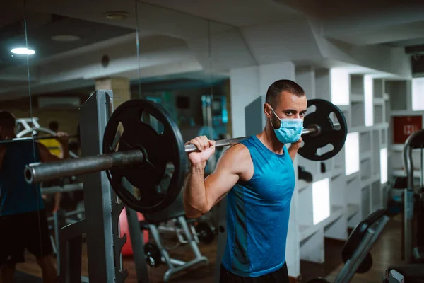 Yüzünde maske olan beyaz bir sporcu spor salonunda ağırlık kaldırıyor ve egzersiz yapıyor. COVID 19 koronavirüs koruması