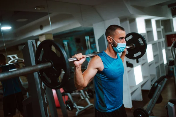 Yüzünde maske olan beyaz bir sporcu spor salonunda ağırlık kaldırıyor ve egzersiz yapıyor. COVID 19 koronavirüs koruması