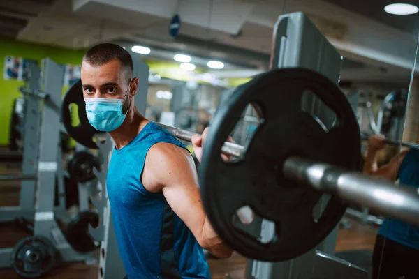 Yüzünde maske olan beyaz bir sporcu spor salonunda ağırlık kaldırıyor ve egzersiz yapıyor. COVID 19 koronavirüs koruması