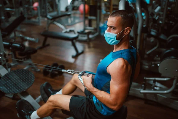 Tıbbi maskeli genç kaslı adam spor salonunda kol ve sırt egzersizi yapıyor. COVID önleme - 19 koronavirüs