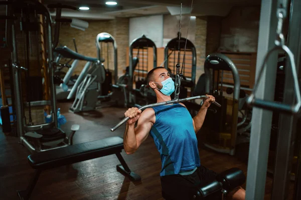 Yüzünde maske olan beyaz bir sporcu spor salonunda ağırlık kaldırıyor ve egzersiz yapıyor. COVID 19 koronavirüs koruması