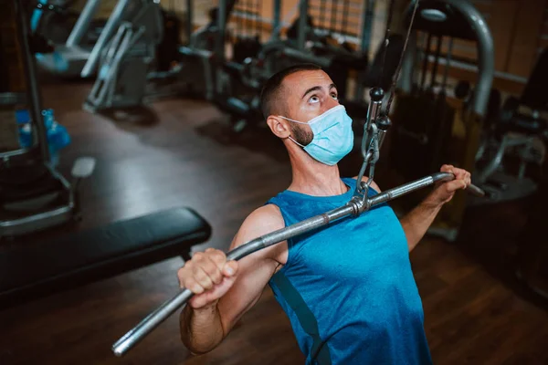 Yüzünde maske olan beyaz bir sporcu spor salonunda ağırlık kaldırıyor ve egzersiz yapıyor. COVID 19 koronavirüs koruması
