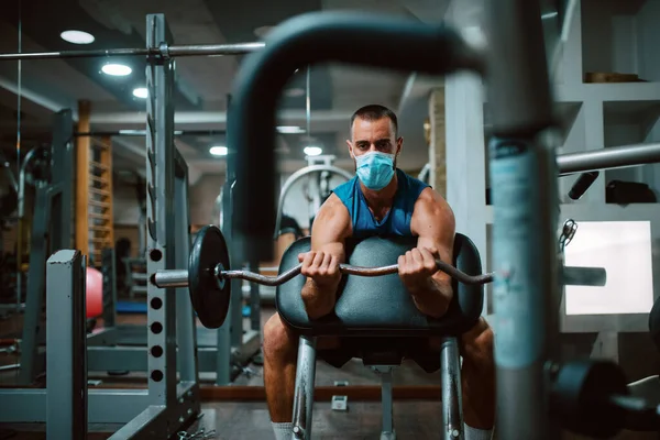 Yüzünde maske olan beyaz bir sporcu spor salonunda ağırlık kaldırıyor ve egzersiz yapıyor. COVID 19 koronavirüs koruması