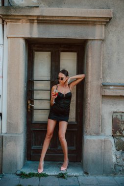 Kapısının önünde elinde kokteyl olan, kısa siyah elbiseli, güzel, çekici, beyaz bir kadın.