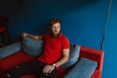 Kırmızı tişörtlü beyaz bir adam kuafördeki bekleme odasında oturmuş dergi okuyor.