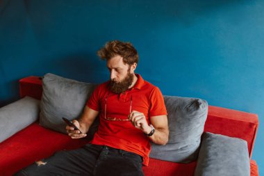 Kırmızı tişörtlü genç bir adam kuaförün bekleme odasında oturuyor ve telefonu kullanıyor.