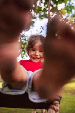 Kırmızı tişörtlü beyaz bir çocuk parkta salıncakta sallanıyor.