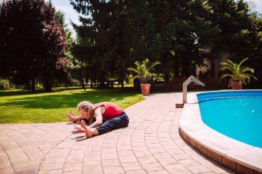 Orta yaşlı, saçları örgülü, beyaz bir kadın havuz kenarında bacaklarını açarak yoga yapıyor.