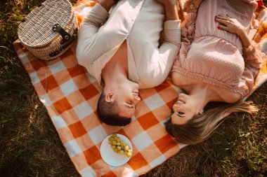 Kafkasyalı bir adam ve sarışın bir kadının piknikte turuncu bir battaniyenin üzerinde yatışının en üst görüntüsü. Yanlarında da bir sepet yiyecek ve üzüm var.