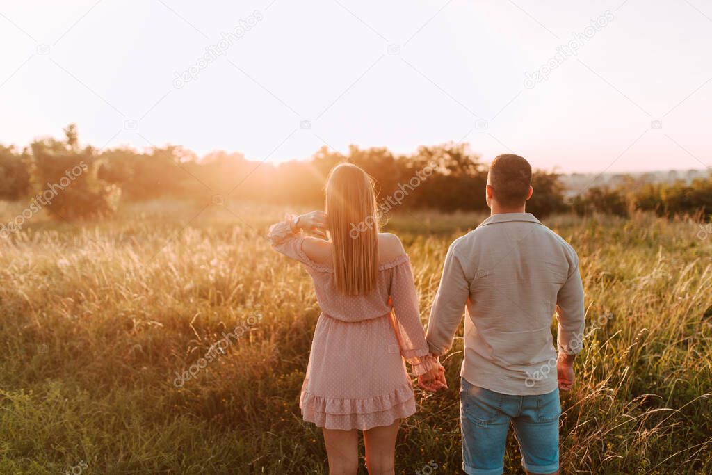 A beautiful blonde caucasian woman in a pastel pink dress and a caucasian man stand in a field at sunset.A loving couple in the nature