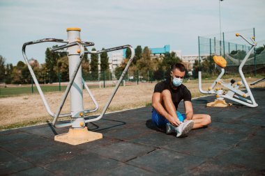 Yüzü maske takan beyaz erkek sporcu, spor salonunda oturur ve bağcıklarını bağlar. Açık havada kaslı bir sporcu. COVID - 19 koronavirüs koruması