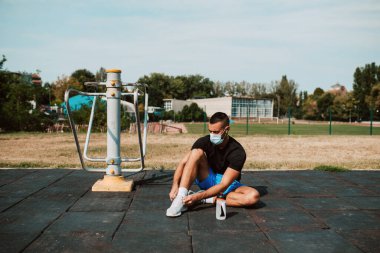 Yüzü maske takan beyaz erkek sporcu, spor salonunda oturur ve bağcıklarını bağlar. Açık havada kaslı bir sporcu. COVID - 19 koronavirüs koruması