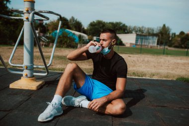 Yüzü maskeli, kaslı beyaz bir erkek sporcu açık hava spor salonunda bir aletin yanında oturur ve şişeden su içer. Coronavirus koruması COVID - 19