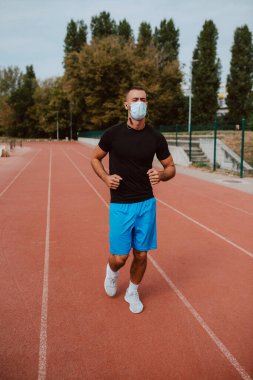 Yarış pistinde yüz maskesi eğitimi almış beyaz bir erkek sporcunun fotoğrafı. Koşucu tüm hava koşullarında atletik koşu pistinde koşar. COVID - 19 koronavirüs koruması