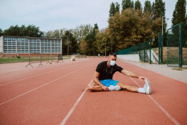 Kulaklıklı, kaslı, beyaz bir erkek sporcu, gözlerini kaçırıyor, stadyum pistinde bacaklarını açıyor. Açık havada kaslı bir sporcu. COVID - 19 koronavirüs koruması