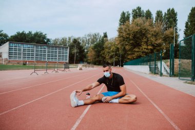Kulaklıklı, kaslı, beyaz bir erkek sporcu, gözlerini kaçırıyor, stadyum pistinde bacaklarını açıyor. Açık havada kaslı bir sporcu. COVID - 19 koronavirüs koruması