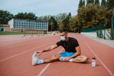 Kulaklıklı, kaslı, beyaz bir erkek sporcu, gözlerini kaçırıyor, stadyum pistinde bacaklarını açıyor. Açık havada kaslı bir sporcu. COVID - 19 koronavirüs koruması