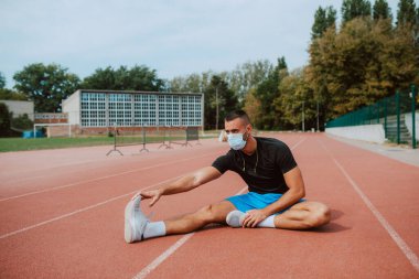Kulaklıklı, kaslı, beyaz bir erkek sporcu, gözlerini kaçırıyor, stadyum pistinde bacaklarını açıyor. Açık havada kaslı bir sporcu. COVID - 19 koronavirüs koruması