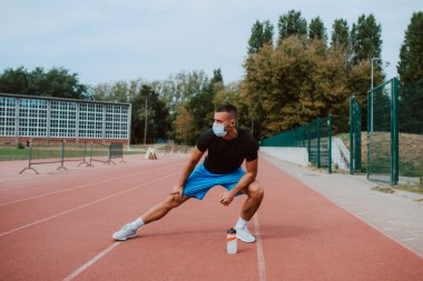 Kulaklıklı, kaslı, beyaz bir erkek sporcu, gözlerini kaçırıyor, stadyum pistinde bacaklarını açıyor. Açık havada kaslı bir sporcu. COVID - 19 koronavirüs koruması