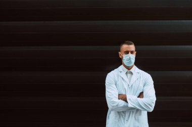 Siyah bir binanın önünde yüzünde koruyucu bir maskeyle beyaz bir doktorun portresi. COVID 'e karşı koruma - 19 koronavirüs