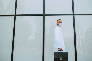 Üniformalı, yüzünde koruyucu bir maske ve kliniğin önünden siyah bir çanta geçen beyaz bir doktor. COVID 'e karşı koruma - 19 koronavirüs