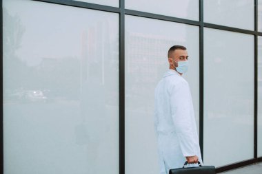 Üniformalı, yüzünde koruyucu bir maske ve kliniğin önünden siyah bir çanta geçen beyaz bir doktor. COVID 'e karşı koruma - 19 koronavirüs