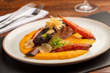 Beyaz bir tabakta patates püresi üzerine sebzeyle süslenmiş dilimlenmiş kaburga ve çatal şarap kadehi ile bulanık bir arka plan.