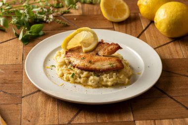 Beyaz tabakta risotto ile kabuklu beyaz tilapia balığı ve limon ve otlarla kaplı bulanık bir arka plan.