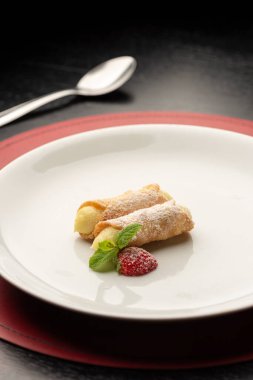 Krema ve çilek ile doldurulmuş Cannoli beyaz tabakta siyah arka planda kaşık ve şarap kadehiyle.