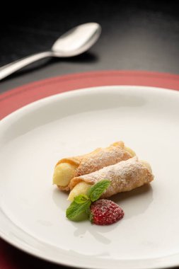 Krema ve çilek ile doldurulmuş Cannoli beyaz tabakta siyah arka planda kaşık ve şarap kadehiyle.