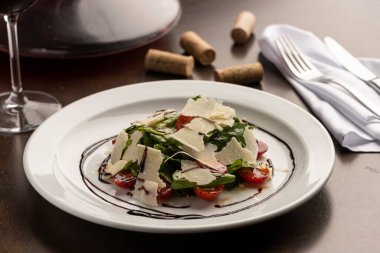 Arugula, domates ve parmesan salatası ahşap bir masa üzerinde balzamik sirke, peçete, şarap şişesi bardağı ve mantarla süslenmiş..