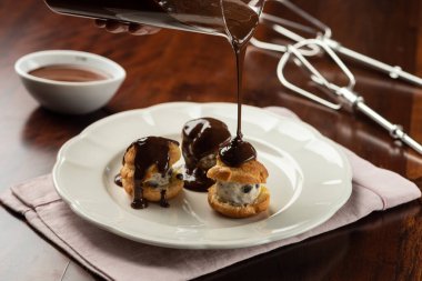 Çikolata kremalı ve kakaolu profiterollü sıcak eritilmiş soslu. Ahşap masadaki arkaplan ve tarif malzemeleri