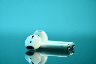 Mavi arka planda beyaz renkli kablosuz kulaklıkların dramatik görüntüsü. Zeminde yansımalar var. Bu ses ve müzik için bir aygıttır