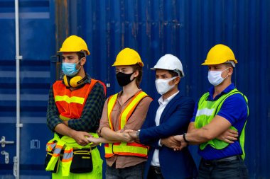 Coronavirus ya da COVID-19 maskeli sanayi işçileri ya da mühendisler kollarını kavuşturup nakliye konteynırının önünde dururlar..