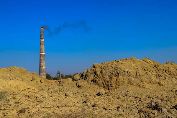 Uzun bir tuğla ocağı bacası gökyüzüne karanlık bir duman yayar, kazılmış toprak yığınlarının arasında durur ve parlak, berrak mavi gökyüzünün altında kırsal bir tel örgü..