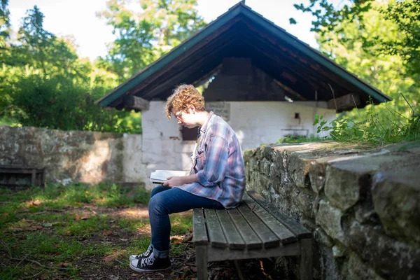 Young woman reads a book on a rustic bench outside. — Stock Photo, Image
