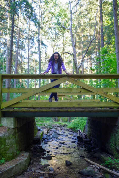 Joven mira a la cámara desde el puente del pie del bosque sobre el arroyo. — Foto de Stock