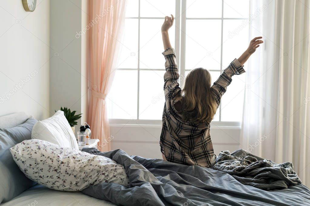 rear view of woman stretching her arms after waking up in morning in front of window