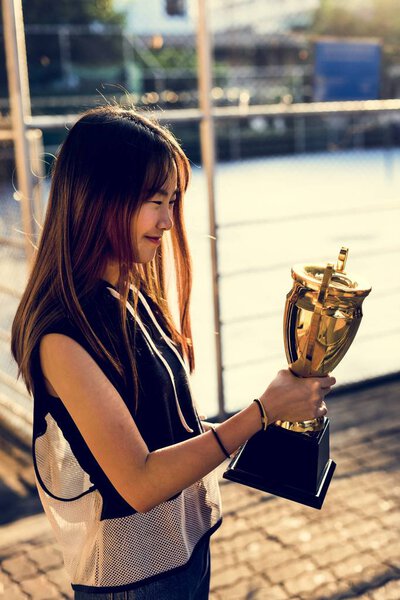 Asian teenage girl in sporty clothes holding a trophy outdoors