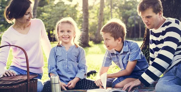 父母和孩子坐在野餐毯子与篮子和书在公园在绿色草 家庭野餐 — 图库照片
