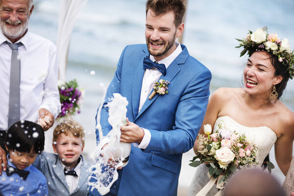 happy young groom opening champagne bottle during wedding ceremony at beach