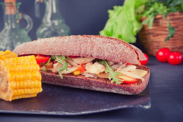 Sanduíche de baguete com peito de peru, queijo, alface, rúcula, tomate e cebola em um prato preto com pedaço de milho . — Fotografia de Stock