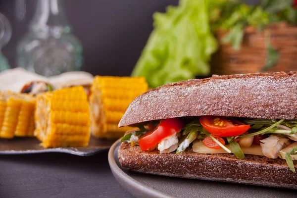 Σάντουιτς μπαγκέτα με στήθος γαλοπούλας, τυρί, μαρούλι, ρόκα, ντομάτα και κρεμμύδι σε ένα μαύρο πιάτο με κομμάτια καλαμποκιού. — Φωτογραφία Αρχείου