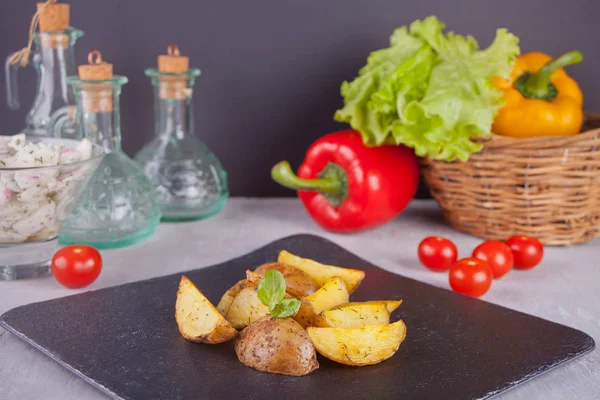 Cuñas de patata horneadas caseras con hierbas en un plato negro con verduras en el fondo —  Fotos de Stock
